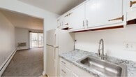 This efficiently designed unit offers four bedrooms, one bathroom, a kitchen, and a connecting hallway, all arranged on a single floor accessible via a staircase. The interior maintains a clean, neutral palette throughout, with bright white walls creating a consistently light and airy atmosphere across all rooms.

The carpeted bedrooms are simply laid out, each featuring baseboard heating and individual wall-unit air conditioners. The spacious primary bedroom includes a sliding glass door opening to a private balcony with views of surrounding trees, providing a notable connection to the outdoors. Additional bedrooms feature windows with natural light and similarly understated finishes.

The kitchen is fitted with white cabinetry, granite-look countertops, a stainless steel sink with a modern faucet, a gas range, and a full-size refrigerator. Light wood-look flooring runs through the kitchen, adding warmth to the space. The hallway connects the kitchen to the rest of the unit seamlessly.

The bathroom features the same light wood-look flooring, a white vanity with under-cabinet storage, and clean, minimal finishes. Overall, the unit presents a straightforward, functional layout with consistent, well-maintained finishes throughout.