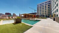 This rooftop amenity level offers a well-rounded collection of indoor and outdoor spaces designed for active living and relaxed social gatherings. The centerpiece is a resort-style swimming pool and adjacent spa, both finished in clean tile and surrounded by a generous concrete deck ideal for sunbathing. Lounge chairs line the pool deck, providing ample space to unwind while taking in panoramic views of the surrounding urban skyline.

The outdoor area extends beyond the pool to a lush artificial turf lounge zone furnished with wooden seating sets and large shade umbrellas, creating a comfortable retreat. A covered pergola with string lighting serves as an al fresco dining area with table-and-chair seating, while a built-in outdoor kitchen with a stainless steel grill provides everything needed for outdoor entertaining. Raised planters with tropical greenery add natural accents throughout the terrace.

The fitness center, housed in a brick-clad structure with expansive floor-to-ceiling glass walls, connects seamlessly to the outdoor deck. Inside, the well-equipped gym features cardio machines, a cable machine, a dumbbell rack, and weight benches, all set on durable rubber flooring. Large mirrors amplify the space, while the glass walls flood the interior with natural light and frame views of the skyline beyond. Exposed ductwork and industrial-style track lighting complete the modern, urban aesthetic.