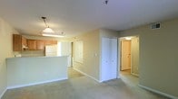 This single-floor residence offers a practical and comfortable layout designed for easy living. The open-concept living area is finished with soft, wall-to-wall carpeting that adds warmth underfoot, while the neutral beige walls create a bright and welcoming ambiance throughout. A peninsula-style kitchen sits adjacent to the living space, featuring wooden cabinetry, a white refrigerator, and overhead lighting including a classic pendant light — making it both functional and visually connected to the main living area. The hallway leading from the living space reveals generous built-in closet storage, with multiple sets of white bi-fold doors offering ample room for organization. A doorway at the end of the hall provides access to additional rooms, suggesting a well-defined separation between living and private spaces. Ceiling-mounted lighting fixtures illuminate each area consistently, and a sprinkler system is visible overhead, reflecting modern safety standards. The overall design is clean and understated, prioritizing functionality with a neutral palette that offers a versatile canvas for personalization. White baseboards and trim throughout add a crisp finish that ties the interior together cohesively.
