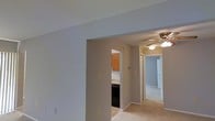This single-floor residential space offers a practical and functional layout designed for comfortable everyday living. The main living area features neutral-toned carpet flooring and light gray walls, creating a clean and understated aesthetic throughout. An arched passageway connects the living space to the kitchen, which is equipped with warm wood-toned cabinetry and dark appliances, providing a classic and efficient cooking environment. A central hallway, illuminated by a ceiling fan with an integrated light fixture, extends toward additional rooms, suggesting a well-organized floor plan with clear separation between living and private spaces. The hallway leads to what appears to be a bedroom or secondary room, visible in the background with its own door for privacy. Soft, neutral wall colors are consistent throughout, contributing to a cohesive and calm interior atmosphere. White baseboards and door trim provide a subtle contrast against the gray walls, adding a touch of definition to the space. The layout flows naturally from the main living area through to the kitchen and private rooms, making the space both accessible and easy to navigate.