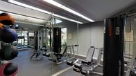 This single-level fitness room is designed to support a well-rounded strength training routine. The space is outfitted with a multi-function cable machine, several adjustable weight benches, a leg press or extension station, and a selection of free weights, providing users with a variety of exercise options. The walls are finished in a neutral gray tone, contributing to a clean and focused environment. Floor-to-ceiling mirrors line one wall, visually expanding the room and allowing users to monitor their form during workouts. Overhead fluorescent lighting ensures the space is consistently well-lit. Rubber flooring throughout offers both durability and safety underfoot. A window along the upper portion of the mirrored wall allows a glimpse of natural light and surrounding greenery, adding a subtle connection to the outdoors. An informational notice posted on the wall suggests the room operates under posted guidelines, indicating shared or community use. Overall, the fitness room presents a practical and functional environment well-suited for regular strength and resistance training.