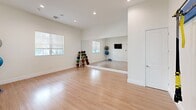 This well-appointed fitness center offers a comprehensive workout experience across a generously sized, single-floor layout. The space is divided into two distinct zones, each designed to support a variety of fitness activities.

The main gym area impresses with soaring vaulted ceilings accented by warm, slatted wood paneling that adds a contemporary and inviting character. Large floor-to-ceiling windows and sliding glass doors flood the space with natural light while offering views of an outdoor patio and pool area. Floor-length mirrors line the walls, visually expanding the room. The gym is outfitted with a full range of cardio equipment — including treadmills, ellipticals, and stationary bikes — as well as a cable machine, free weights, kettlebells, and a dumbbell rack. Teal and neutral-toned carpet tiles define the exercise zones, and ceiling fans and linear pendant lighting provide comfort and visibility throughout.

The secondary studio room features light hardwood flooring, recessed lighting, and a clean white palette that creates a calm, open atmosphere ideal for yoga, stretching, or functional training. This room is equipped with TRX suspension trainers, medicine balls, stability balls, wall-mounted mirrors, and a mounted display screen, making it a versatile space for group classes or individual training sessions.