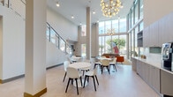 Located at 787 NW 43rd Ave in Miami, FL, this single-story commercial space presents a striking, contemporary design centered around a grand, double-height lobby. Expansive floor-to-ceiling windows line the exterior-facing walls, flooding the interior with abundant natural light and offering open views of the surrounding streetscape and landscaping. The lofty ceilings create an airy, voluminous atmosphere enhanced by modern sputnik-style chandeliers that serve as decorative focal points.

The reception area features a sleek, sculptural desk with a dark wood-paneled base and a crisp white countertop, set against a dramatic floor-to-ceiling polished marble accent wall in rich brown and grey tones. Adjacent frosted glass partitions provide a refined separation of spaces while maintaining visual openness. Vibrant, large-scale abstract artwork punctuates the neutral walls, adding energy and color throughout.

A furnished lounge area with contemporary white chairs, round tables, and colorful area rugs invites gathering and informal interaction. Glazed double doors lead to a well-equipped fitness center visible from the main corridor. Light-toned large-format tile flooring and a staircase with cable-rail detailing complete the polished, professional aesthetic of this thoughtfully designed space.