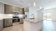 This well-appointed 1-bedroom, 1-bathroom residence offers a thoughtfully designed open-concept layout that maximizes functionality and style. The combined kitchen and living area serves as the heart of the home, featuring light wood-tone flooring, recessed lighting, and a ceiling fan. The kitchen is outfitted with two-tone cabinetry in warm wood finishes, a granite-topped island with an undermount sink and pendant lighting overhead, white subway tile backsplash, and a full suite of stainless steel appliances. A built-in nook near the entry provides convenient storage with hooks and shelving. The living area is bright and airy, enhanced by a sliding glass door that opens to a private balcony, inviting ample natural light. The bedroom features plush carpeting, a ceiling fan, and a window with views of the surrounding area. A dedicated closet offers additional storage. The bathroom is clean and contemporary, with large-format tile flooring, a wood-toned vanity with granite countertop, and a glass-enclosed walk-in shower with white tile surround. A hallway connects the spaces efficiently, completing this cohesive and modern floor plan.