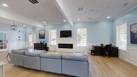 This single-story clubhouse in Horn Lake, Mississippi offers a thoughtfully designed community gathering space with a bright, welcoming atmosphere throughout. The interior is unified by soft powder-blue walls, crisp white wainscoting, and light hardwood flooring that flows seamlessly from room to room.

The centerpiece of the clubhouse is a spacious open lounge area, anchored by a sleek linear fireplace set within a white shiplap surround, with a wall-mounted flat-screen TV positioned above. A generous sectional sofa invites relaxed gathering, while recessed lighting and multiple white-shuttered windows fill the space with abundant natural light. Structural columns with coffered ceiling details add architectural character and define distinct zones within the open floor plan.

A front reception and workspace area provides a functional desk station, ideal for administrative use or resident services. The adjoining kitchen is fully equipped with stainless steel appliances, white shaker cabinetry, a marble-style tile backsplash, and a peninsula with bar seating — making it well-suited for hosting events and social gatherings. Large windows in the kitchen frame views of the surrounding greenery.

A corridor leading through the space features vaulted ceiling accents, decorative artwork, and elegant pendant lighting, lending the clubhouse a polished, inviting character. French doors provide access to the exterior, enhancing the sense of openness and connectivity to the outdoors.