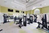 Located at 9800 Touchton Rd in Jacksonville, FL, this fitness center offers a spacious, single-floor workout facility designed to support a comprehensive exercise experience. The room is distinguished by soaring tray and vaulted ceilings accented with recessed lighting and a large ceiling fan, creating an open, airy atmosphere. Walls painted in a fresh sage green with white wainscoting lend the space a clean, energizing aesthetic.

Natural light floods the room through an array of large multi-pane windows and a prominent arched transom window, providing views of the surrounding greenery. Full-length mirrors line one wall, amplifying the sense of space and allowing users to monitor their form.

The facility is comprehensively equipped for both strength and cardiovascular training. The strength zone features a full dumbbell rack, adjustable benches, a power rack, and multiple Freemotion cable machines. The cardio area includes treadmills and additional cardio equipment positioned near the windows. Multiple wall-mounted flat-screen TVs are positioned throughout for entertainment during workouts.

The flooring consists of neutral carpet tiles with yellow-green accent squares, adding a modern touch. An adjacent small room with hardwood flooring provides additional utility space, rounding out this well-appointed fitness facility.