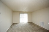 This single-floor residential unit offers a straightforward and functional layout designed for comfortable everyday living. The living room is generously sized, featuring neutral-toned wall-to-wall carpeting, a textured ceiling, and a large sliding glass door fitted with vertical blinds that allows abundant natural light to fill the space. An entry door with a light switch panel completes the room.

The kitchen is positioned in an open-concept arrangement, accessible via a pass-through counter that connects it to the living area. It is equipped with a white electric range, a range hood, and warm-toned wood cabinetry providing ample upper and lower storage. A double sink is integrated into the counter, and track lighting overhead ensures the workspace is well-lit. The kitchen floor features wood-look vinyl, contrasting with the carpeted living area.

The bedroom is a comfortable, carpeted room with neutral walls and a textured ceiling. A sliding closet with organized rod storage provides practical wardrobe space, and a window offers natural light and an exterior view.

The bathroom features wood-look vinyl flooring and includes a long vanity with a single sink, a large mirror with overhead lighting, and a toilet. A towel bar and additional door access provide convenience and privacy. Overall, the unit presents a clean, functional design with consistent neutral finishes throughout.