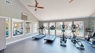 Located at 16200 Space Center Blvd in Houston, TX, this fitness center offers a well-organized and thoughtfully designed workout facility spread across a single floor. The space is divided into two distinct areas, each serving a specific fitness purpose.

The main cardio room features vaulted ceilings with a ceiling fan, recessed lighting, and large windows that frame views of the outdoor pool and patio area. Durable rubber flooring runs throughout, and full-length mirrors along one wall amplify the sense of space. The room is equipped with multiple treadmills, elliptical trainers, and stationary bikes, providing a comprehensive cardio workout experience.

The secondary room offers a dedicated strength and flexibility zone, housing a rack of dumbbells in varying weights alongside stability balls and medicine balls for functional training. Natural light pours in through multiple windows overlooking lush tropical landscaping, creating a bright and energizing environment.

A tiled entryway connects both areas, providing a clean transition between zones. Neutral taupe walls, white crown molding, and abundant windows throughout contribute to an open, airy ambiance. The poolside orientation of the facility enhances the overall experience, offering motivating outdoor views during every workout.