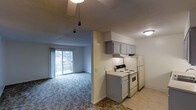 This single-floor space offers a functional and open layout that seamlessly connects the kitchen and living area. The kitchen is equipped with white appliances, including an electric range and refrigerator, and features gray cabinetry with an overhead range hood, providing generous storage. The tile flooring in the kitchen transitions into plush carpeting in the adjoining living area, creating a clear yet cohesive distinction between the two spaces. A pendant light fixture adds a decorative touch to the open-concept layout. The living area is bright and spacious, benefiting from a large sliding glass door that opens onto a private balcony, allowing abundant natural light to fill the room. The balcony offers an outdoor retreat with views of the surrounding area. Neutral wall tones and a textured ceiling throughout contribute to a clean and understated aesthetic. The overall design prioritizes practicality and comfort, making it a well-organized and inviting single-floor space suited for everyday living.