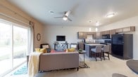 This two-floor residence showcases a thoughtfully designed open-concept main level where the living, dining, and kitchen areas flow together without visual interruption. The living area is anchored by a ceiling fan with integrated lighting and features light hardwood-style flooring that extends throughout the space, contributing to a bright and cohesive atmosphere. Large windows dressed with curtains allow natural light to filter into the room, enhancing the airy feel of the layout.

The kitchen is positioned seamlessly adjacent to the living space and is outfitted with dark espresso cabinetry, dark countertops, and black appliances. A kitchen island with bar-height seating serves as both a functional workspace and a natural gathering point. Pendant lights hang above the island, adding focused task lighting and a touch of visual interest.

The overall color palette throughout the main level is neutral and contemporary, combining warm grey tones with dark wood accents. The open layout and connected design of the living and kitchen areas make this space well-suited for everyday living and entertaining alike, while the second floor provides additional private living space above.