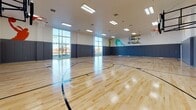 The Devon's indoor basketball court is a well-appointed, full-sized athletic facility designed to accommodate serious recreational play. The court features a polished hardwood floor with clearly marked regulation lines for multiple court configurations, allowing for a variety of games and drills simultaneously. Multiple basketball hoops are positioned at each end of the court, providing ample playing options for individuals and groups alike.

The perimeter walls are lined with padded blue panels extending to mid-wall height, offering both a protective barrier and a clean, professional aesthetic. Above the padding, the white upper walls are adorned with bold, dynamic silhouette murals of basketball players in various athletic poses, rendered in gold, teal, and yellow tones, adding energy and character to the space.

The soaring ceiling height contributes to an open, airy atmosphere, while rows of recessed rectangular fluorescent fixtures provide consistent, bright illumination across the entire playing surface. Large floor-to-ceiling windows along one wall allow natural light to flood the court and offer views of the surrounding exterior, enhancing the overall sense of openness. Access doors are integrated discreetly into the padded wall sections, maintaining the facility's streamlined appearance.