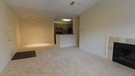 This single-floor residence offers an open and functional layout designed for comfortable everyday living. The main living area is spacious and airy, finished with light-toned carpeting that extends throughout the space, creating a warm and cohesive feel. Neutral beige walls and standard-height ceilings contribute to a clean, understated aesthetic.

The kitchen is positioned at the heart of the home and features medium-toned wood cabinetry, dark countertops, and a peninsula-style breakfast bar that serves as a natural divider between the kitchen and living area. Black appliances, including a range and refrigerator, are integrated into the cabinetry, and overhead lighting provides ample illumination for meal preparation.

The living area is generously sized and benefits from track lighting, offering flexibility in how the space is lit and used. The open-plan design between the kitchen and living room encourages a connected and social atmosphere. Visible HVAC vents in the ceiling indicate a centralized climate control system. Clean white baseboards frame the carpeted floors throughout, adding a crisp finishing detail. Overall, the space presents a practical and move-in ready environment with a neutral palette that allows for a variety of interior design possibilities.