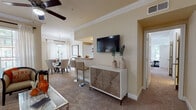This single-story residence offers a thoughtfully designed open-concept layout that seamlessly connects the living, dining, and kitchen areas. The living room is finished with plush carpeting, crown molding, and a ceiling fan, while large windows dressed with white blinds and sheer curtains allow generous natural light to fill the space. A wall-mounted television and a stylish storage console anchor the room, complemented by a breakfast bar with barstool seating that bridges the living area to the kitchen.

The kitchen features white cabinetry with wood-trimmed accents, tile flooring, ample counter space, and a full suite of white appliances including a range, microwave, refrigerator, and dishwasher. The dedicated dining area accommodates a round glass-top table beneath a pendant light fixture, with a window providing an airy, well-lit atmosphere.

The bedroom is carpeted and bright, with natural light filtering through blinds and access to a spacious en-suite bathroom. The bathroom offers an expansive vanity with dual countertop areas, recessed lighting, and tile flooring. A dedicated laundry closet with a full-size washer and dryer adds everyday convenience, rounding out the functional layout of this residence.