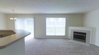 This two-floor residence offers a functional and comfortable layout designed to maximize natural light and livability. The main living area is a central highlight, featuring wall-to-wall carpeting in a neutral tone that complements the soft, beige wall color throughout. A large picture window and an adjacent sliding glass door fitted with vertical blinds flood the room with abundant natural light while providing views of the surrounding outdoor greenery. The textured ceiling adds a classic architectural detail consistent throughout the space.

A standout feature of the living area is the tile-surround fireplace positioned along one wall, offering both a practical heating source and a focal point for the room. The open sightlines from the living space toward what appears to be a kitchen counter suggest an open-concept flow between rooms, enhancing the sense of spaciousness. The neutral color palette of beige walls and light carpet creates a versatile backdrop suitable for a variety of interior styles. Spanning two floors, the residence provides a clear separation between living and private spaces, adding to the overall functional appeal of the home.