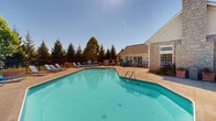 Located at 437 Sunburst Dr in Delaware, Ohio, this outdoor amenity area is centered around a generously sized swimming pool with clear, turquoise water. The pool features convenient entry via stainless steel handrails and is surrounded by a broad concrete sun deck lined with blue lounge chairs, providing ample space for sunbathing and relaxation.

Adjacent to the pool, an outdoor patio area is outfitted with high-top tables and blue chairs, ideal for casual outdoor dining or socializing. A full-size gas grill station is conveniently positioned nearby, making the space well-suited for outdoor gatherings and cookouts.

The area is enclosed by a black wrought iron fence that provides both security and an open sightline to the surrounding mature trees and landscaping, which offer a natural, private backdrop. A single-story clubhouse or amenity building with stone and vinyl siding anchors one side of the pool area, featuring large windows that overlook the deck. A stone chimney adds a classic architectural touch to the structure.

The overall layout is thoughtfully designed to maximize outdoor enjoyment, with the pool, lounging, dining, and grilling areas all within close proximity. Under open skies and surrounded by lush greenery, this outdoor space offers a welcoming and functional recreational environment for residents.