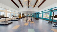 Located at 633B SW 150th St in Burien, WA, this fitness center offers a spacious, contemporary workout environment designed to accommodate a wide range of exercise needs. The main workout area is architecturally distinguished by soaring ceilings with rich, dark exposed wood beams and striking circular pendant lighting that creates a dynamic, energetic atmosphere. Large windows line the walls, flooding the space with natural light and providing views of the surrounding exterior.

The flooring throughout the main gym features a durable, multi-toned rubber surface with colorful inlaid sections, offering both safety and style. The cardio zone is well-stocked with treadmills, stair climbers, stationary bikes, and ellipticals, complemented by wall-mounted televisions and a vibrant nature-themed mural along one wall. A dedicated strength training area includes a power rack with Olympic weight plates, cable machines, a full dumbbell rack, and benches, all set against bold teal accent walls with large mirrors.

A separate studio room features light hardwood flooring, linear ceiling lighting, and open wall space, making it ideal for group classes or functional training, with storage for medicine balls and fitness accessories. An accessible restroom and water station are also available. The overall design balances high energy with functionality, creating an inviting and comprehensive fitness facility.