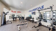 Located at 101 Barnes Lane in Broussard, Louisiana, this fitness center delivers a fully equipped workout environment designed to inspire and perform. The single-floor space is finished with durable rubber flooring throughout, white walls, and recessed ceiling lighting that keeps the atmosphere bright and energizing. Bold motivational phrases — "Train Hard," "It's a Lifestyle," and "Feel the Burn" — are displayed prominently across the walls, reinforcing the space's commitment to fitness culture. Large floor-to-ceiling windows and glass entry doors flood the interior with natural light and offer views of the surrounding outdoors, enhancing the open, airy feel of the facility. Full-length wall mirrors are strategically placed to maximize visibility and give the space a larger, more expansive appearance. The equipment layout is thoughtfully organized, featuring a dedicated free weights area with a multi-tiered dumbbell rack, adjustable benches, a cable crossover machine, and a functional trainer. Strength machines for targeted muscle groups line the perimeter, while cardio equipment including treadmills and stationary bikes occupy additional floor space. A wall-mounted flat-screen TV is also present for entertainment during workouts. The facility is well-ventilated and finished to a high standard throughout.
