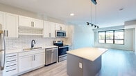 This well-designed single-floor unit offers a thoughtfully laid-out floor plan encompassing a kitchen, two living areas, and two bathrooms. The open-concept kitchen is a standout feature, showcasing white shaker-style cabinetry with matte black hardware, a classic white subway tile backsplash, and a full suite of stainless steel appliances including a refrigerator, range, microwave, and dishwasher. A central gray island with a white quartz countertop provides additional workspace and seating, while modern pendant and track lighting add a stylish finishing touch. Light wood-style flooring flows seamlessly throughout the entire unit, enhancing the bright and airy atmosphere. The living areas benefit from large black-framed windows that invite abundant natural light and offer outdoor views, with one area featuring direct access to a private balcony through a sleek black-framed glass door. The bathrooms are finished with white cabinetry, quartz countertops, and matte black fixtures, with one bathroom conveniently housing a stacked washer and dryer unit. A walk-in closet with built-in shelving adds practical storage. Clean white walls and recessed lighting throughout contribute to the unit's fresh, contemporary aesthetic.