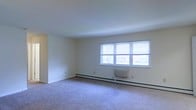 This single-floor space features a generously sized main living area finished with wall-to-wall carpet flooring in a neutral tone, contributing to a clean and understated aesthetic. The room is characterized by its bright, airy atmosphere, largely due to a set of double windows along the exterior wall that allow ample natural light to fill the space. The windows are fitted with horizontal blinds, offering privacy and light control as needed. The walls are painted in a crisp, neutral white, enhancing the sense of openness throughout. Heating is provided by a baseboard heating system that runs along the base of the exterior wall, complemented by a wall-mounted unit positioned beneath the windows — practical features well-suited for year-round comfort. The smooth, white ceiling maintains a clean overhead plane that keeps the space feeling open. An open entryway or passageway is visible along one wall, suggesting connectivity to additional areas of the unit. Overall, the space presents a simple, functional layout with a neutral palette that offers a versatile foundation for a variety of interior configurations.