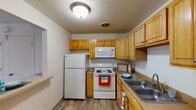 This single-story residence offers a practical and comfortable layout across several well-defined living spaces. The kitchen is efficiently designed with warm honey-toned wood cabinetry extending to the upper walls, providing generous storage throughout. White appliances, including a refrigerator, electric range, and over-the-range microwave, are neatly integrated alongside a double-basin stainless steel sink. Wood-look flooring adds a clean, durable finish to the space. A pass-through counter connects the kitchen to the living area, enhancing the open feel between the two spaces.

The living area is generously sized, featuring neutral carpet, bright white walls, and a ceiling fan overhead. Large sliding glass doors lead to an outdoor patio, allowing natural light to fill the room and providing views of the surrounding greenery. A separate entry door is also located in this space.

The bedroom is a quiet, carpeted room with a large window dressed in vertical blinds, offering ample natural light. A closet provides convenient storage.

The bathroom is straightforward and clean, with wood-look flooring, white walls, and a full tub-shower combination framed by a decorative curtain. The layout is efficient and well-lit, completing this functional living space.