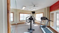 Located in St. Andrews, this dedicated fitness room is thoughtfully designed to support a well-rounded workout routine. The single-floor space is finished with neutral, warm-toned walls and carpeted flooring, creating a comfortable and inviting environment for exercise. Large windows framed in warm wood trim line the walls, flooding the room with natural light and offering views of the surrounding outdoor area.

The room is equipped with a selection of cardio machines, including a treadmill and an elliptical trainer, as well as a multi-station cable weight system suitable for a variety of strength training exercises. A medicine ball and additional accessories are also available. A wall-mounted flat-screen television provides entertainment during workouts, while a ceiling fan ensures comfortable air circulation throughout the space.

A full-length wall mirror spans one side of the room, enhancing the sense of openness and allowing users to monitor their form during exercise. French doors on one end of the room provide access to an adjacent exterior area, further brightening the space. A bold red accent wall bearing the motivational phrase "Pursue Excellence" adds a dynamic, energizing touch to the overall design, reinforcing the room's purpose as a space dedicated to health and fitness.
