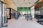 Located at 3461 La Cadena Dr in Riverside, CA, this well-appointed fitness center spans a single floor and offers a comprehensive workout environment across two distinct areas. The main gym floor is generously sized with soaring ceilings, recessed lighting, and large floor-to-ceiling windows that flood the space with natural light and provide views of the surrounding exterior. A bold green accent wall anchors the back of the room, adding a vibrant, energizing aesthetic to the neutral palette. An overhead ceiling fan complements the open, airy feel of the space. The main floor is outfitted with a full row of treadmills and stair climbers, multiple weight benches, a True brand cable and functional trainer system, a dumbbell rack, and additional strength machines. Glass-panel partition walls separate the main gym from an adjacent enclosed studio, which features warm wood-look flooring, floor-to-ceiling mirrors, spin bikes, stability balls, and a dedicated TRX and functional training rig complete with a punching bag, kettlebells, and suspension trainers. Together, these two zones create a versatile, full-featured fitness facility suited for cardio, strength, and group or individual training.