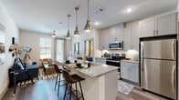 This thoughtfully designed single-floor residence combines contemporary finishes with a functional open-concept layout. The kitchen serves as the heart of the home, showcasing light gray shaker-style cabinetry, granite countertops, a stainless steel appliance suite including a range, microwave, and refrigerator, and a herringbone tile backsplash. A central island with seating for two is elegantly lit by three industrial-style pendant lights, making it ideal for casual dining and entertaining. Wood-look plank flooring flows seamlessly throughout the space, connecting the kitchen to an open living area bathed in natural light from large windows fitted with horizontal blinds. The bedroom is a serene retreat, featuring generous windows with gold-toned curtain panels and warm wood furniture accents. The bathroom is well-proportioned, offering a granite-topped vanity with ample cabinetry, a glass-enclosed walk-in shower, and access to a dedicated walk-in closet. A separate in-unit laundry area houses a full-size stacked washer and dryer for added convenience. Throughout the residence, a neutral color palette with warm accent tones creates a cohesive, modern aesthetic.