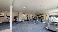 Located at 175 S Rio Vista St in Anaheim, California, this fitness center offers a practical and functional space designed to support a well-rounded workout routine. The single-floor layout is divided into two areas: a spacious exercise room and a dedicated bathroom.

The exercise room is the heart of the facility, finished with durable blue-gray carpet tiles that provide comfortable underfoot support during workouts. Recessed lighting illuminates the space evenly, while a large wall of floor-to-ceiling mirrors along one side visually expands the room and allows users to monitor their form. The equipment selection includes stationary bikes, an elliptical cross-trainer, a rowing machine, a treadmill, and a multi-station strength training machine, offering a comprehensive range of cardio and resistance options. Framed artwork adorns the neutral-toned walls, adding a touch of warmth to the utilitarian setting. A window near the entrance allows natural light to filter in, and a keyed entry door provides secure access directly from the exterior.

The bathroom is a clean, no-frills facility featuring tile flooring, a toilet, a mounted paper towel dispenser, and a soap dispenser — providing essential post-workout convenience. Together, the two spaces form a compact yet complete fitness amenity well-suited for regular use.
