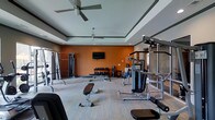The fitness center is a spacious, single-level facility designed to accommodate a comprehensive workout experience. The room features a tray ceiling with recessed lighting and dual ceiling fans, ensuring proper ventilation and a well-lit environment throughout. Bold orange accent walls add an energetic atmosphere, contrasting with the neutral gray tones of the ceiling and flooring.

The space is outfitted with a wide selection of strength-training equipment, including a multi-station cable machine, adjustable weight benches, and a full dumbbell rack offering a range of free weights. Cardio and functional training machines are also present, providing options for varied workout routines. The open floor plan allows for comfortable movement between stations without congestion.

A wall-mounted flat-screen television is positioned prominently for entertainment during workouts. Large glass panels or windows along one side of the room allow natural light to filter in, contributing to an open and airy feel. The carpeted flooring provides traction and comfort underfoot. Overall, the fitness center is thoughtfully arranged to support both strength and general fitness goals in a clean, modern setting.