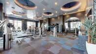 The Elysian Fitness Center is a thoughtfully designed, upscale fitness facility that combines high-end aesthetics with a comprehensive array of exercise equipment. The space is entered through a stylish vestibule featuring large glass doors, an "Elysian" branded welcome mat, and a decorative water feature wall accented by tropical greenery, setting an inviting tone from the moment of arrival.

The main workout floor is expansive and open, finished with a distinctive patterned carpet in layered tones of blue, brown, and grey that adds visual character throughout. Dark charcoal accent walls contrast elegantly with the light-toned ceiling, which is adorned with multiple circular tray insets housing ceiling fans and ambient lighting — a standout architectural feature that elevates the space.

Large windows along the perimeter flood the interior with natural light and provide views to the surrounding outdoor environment. Oversized round wall mirrors are strategically placed to enhance depth and visibility across the floor.

Equipment is organized into clearly defined zones, including a cardio section with treadmills, ellipticals, and stationary bikes from TRUE and other premium brands, a dedicated strength training area with a full rack of dumbbells and weight benches, and a selectorized machine circuit for targeted muscle training. A functional training station with a cable machine and free weights completes the comprehensive offering.