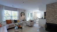 This single-floor residence offers an open-concept layout designed to maximize both functionality and visual appeal. The living area serves as the centerpiece of the space, featuring neutral walls and smooth ceilings that create a clean, airy atmosphere. Large windows dressed with floor-length white curtains flood the room with natural light, offering views of surrounding greenery and enhancing the sense of openness throughout.

The living area is generously proportioned, comfortably accommodating a full sofa and lounge seating arrangement centered around a round glass coffee table. Decorative touches, including a large octagonal wall clock and sculptural accents, contribute character without overwhelming the understated aesthetic.

The space transitions seamlessly into a dining area and a kitchen featuring a dark-topped breakfast bar with counter seating — ideal for casual dining or entertaining. A recessed ceiling detail marks the transition between spaces, adding subtle architectural interest.

Throughout, light-toned flooring and a plush area rug anchor the living zone, while consistent use of soft whites and greys creates a cohesive, calming palette. The overall ambiance is welcoming, elegant, and well-suited for everyday living.