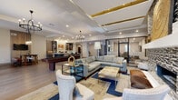 This elegantly designed single-floor communal space blends contemporary style with warm, inviting elements across a generous open-concept layout. Wide-plank hardwood flooring flows throughout the main areas, unifying the various functional zones with a cohesive, polished aesthetic.

The expansive lounge area serves as the heart of the space, anchored by a striking floor-to-ceiling mosaic tile fireplace wall that commands immediate attention. Comfortable sectional seating, layered area rugs in blue and neutral tones, and glass-topped coffee tables create an atmosphere ideal for gathering and relaxation. Overhead, a coffered ceiling with gold accent inlays and a statement chandelier add architectural drama.

Adjacent to the lounge, a spacious kitchenette and bar area features sleek white countertops, dark wood cabinetry, open shelving, and a beverage station — designed to support both casual use and hosted events. A central island with bar seating provides additional workspace and social interaction.

A dedicated dining nook offers a rustic-modern contrast, with a substantial wood and metal table, tufted upholstered chairs, pendant lighting, and a wood-paneled accent wall with a mounted television. Large windows dressed in floor-length drapes fill the area with natural light.

A corridor with large-format stone tile flooring leads to private office or meeting rooms enclosed with frosted glass doors, providing quiet, functional spaces. The overall ambiance is upscale, warm, and professionally curated.
