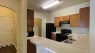 This single-floor space is thoughtfully designed to maximize functionality and comfort. The kitchen serves as a central highlight, featuring warm honey-toned wood cabinetry that provides generous storage throughout. A full suite of black appliances — including a refrigerator, electric range, and built-in range hood — equips the space for everyday cooking needs. The countertops offer a speckled, light-toned surface that extends into a convenient breakfast bar, ideal for casual dining or additional workspace. A stainless steel sink is positioned along the counter, adding a practical finishing touch. The kitchen's layout follows an efficient L-shape, allowing for easy movement and workflow. Overhead, a bright fluorescent fixture ensures the workspace is well-lit. A doorway visible from the kitchen connects to an adjacent room, suggesting an open and accessible floor plan. The walls are finished in a neutral tone that complements the cabinetry, while the textured ceiling adds subtle visual depth. Overall, the space presents a clean, utilitarian design with a welcoming atmosphere suited for everyday living.
