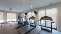 Located at 3443 Park St in Grove City, Ohio, this well-appointed fitness center offers a comprehensive single-level layout designed to support a full range of workout routines. The cardio area is equipped with multiple treadmills, elliptical machines, and a rowing machine, all positioned along large windows that flood the space with natural light and provide views of the surrounding neighborhood. Wall-mounted televisions are conveniently placed throughout, and a ceiling fan overhead keeps the area comfortable during intense sessions. A striking black-and-white athletic mural adds a motivational aesthetic to the cardio zone. An adjoining free weights section features a full rack of dumbbells in varying sizes, a workout bench, and floor-to-ceiling mirrors, creating a practical and spacious strength training environment. A separate studio room provides an open, hardwood-style floor ideal for group classes or solo training, complete with TRX suspension straps, a large display screen with interactive workout programming, and motivational signage. The transition area between zones includes a SmartMat yoga guide, medicine balls, and additional accessories for stretching and warm-up. Recessed lighting, neutral tones, and clean sightlines throughout give the facility a modern, professional atmosphere well-suited for daily use.