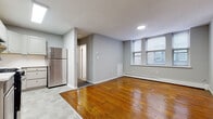 This single-floor unit is characterized by warm hardwood flooring that runs throughout the main living areas, complemented by cool gray walls that lend a clean, neutral backdrop to the space. The open living area benefits from natural light streaming through a set of triple windows fitted with horizontal blinds, offering views of the surrounding exterior. A baseboard heating system runs beneath the windows, ensuring consistent warmth throughout. The kitchen is a well-defined area, transitioning from hardwood to a geometric hex-patterned tile floor. It features gray cabinetry with ample upper and lower storage, light countertops, and a full suite of stainless steel appliances including a gas range, overhead ventilation hood, and a full-size refrigerator. A utility sink completes the functional layout. The unit extends into additional rooms connected by doorways, with the floor plan revealing a series of interconnected spaces, including what appears to be a bedroom and a separate carpeted room. Recessed and flush-mount lighting fixtures illuminate the interior, while white-painted trim and baseboards provide clean contrast throughout. The overall layout is practical and straightforward, offering a functional and livable environment.