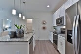 This single-story residence is designed with a clean, contemporary aesthetic throughout, combining neutral tones, light wood-style flooring, and plush carpeting in the bedrooms. The open-concept kitchen is a standout feature, equipped with stainless steel appliances, a built-in microwave, white shaker-style cabinetry, granite countertops, and a tile backsplash. A central island with seating provides additional workspace and flows seamlessly into the dining area, which is illuminated by a trio of pendant lights overhead. The living room offers a comfortable, airy atmosphere with ceiling fan, large windows with blinds and drapes, and direct access to an outdoor space. The primary bedroom is generously sized with plush carpet, a ceiling fan, and direct access to an en-suite bathroom featuring a granite vanity, glass-enclosed shower, and a walk-in closet. A second full bathroom includes a white vanity with granite countertop and a tub-shower combo. A dedicated laundry area with wire shelving adds everyday convenience. The hallway features light wood flooring and connects each room with a cohesive, well-organized layout throughout.
