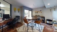 Located at 840 17th St in San Diego, CA, this single-floor space presents a well-appointed, modern interior with an industrial-meets-contemporary aesthetic. The main gathering area is anchored by a round dark wood dining table surrounded by cognac leather chairs, all set atop a plush shag area rug over warm terracotta tile flooring. The space features a combination of recessed lighting and distinctive pendant fixtures, including an eye-catching pulley-style floor lamp that adds an industrial character to the room.

A long wooden counter along one wall provides additional seating with adjustable bar stools, while wall-mounted artwork in warm yellows and grays adds a pop of color against the neutral gray and slate-toned walls. Large glass sliding doors and floor-to-ceiling windows invite abundant natural light and offer direct access to an outdoor patio area framed by decorative iron railings and lush greenery.

An interior glass partition wall separates a secondary workspace or office area, which is visible from the main gathering space and is equipped with office equipment including a printer. The open layout and thoughtful furnishings make this a versatile, functional space well-suited for meetings, community gatherings, or collaborative work in the heart of San Diego.