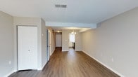 This two-floor space offers a clean, functional layout with a cohesive design throughout. The main level features warm hardwood-style flooring that extends across the open living and dining areas, complementing the neutral, light gray wall tones and bright white trim and baseboards. A textured ceiling with a central HVAC vent contributes to the practical, well-maintained character of the space.

The living area flows naturally toward a defined dining zone, distinguished by a structural beam overhead and a classic chandelier that adds a subtle decorative touch. Beyond the dining area, the kitchen is partially visible, featuring cabinetry, a window that allows natural light to filter through, and standard appliances including a range.

A hallway connects the main living spaces to additional rooms, with a staircase leading to the second floor. A doorway near the entry provides access to a closet or additional room, enhancing storage options on the main level. The overall ambiance is bright, neutral, and move-in ready, with consistent finishes that lend the space a tidy, modern feel suited to a variety of living arrangements.