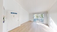 This well-appointed 1-bedroom, 1-bathroom single-story unit offers a clean, modern aesthetic throughout, characterized by a crisp white palette, light hardwood flooring, and an abundance of natural light. The combined kitchen and living room serves as the heart of the unit. The living area features notably high ceilings accented by elegant crown molding and a ceiling fan, creating an airy, open ambiance. Large sliding glass doors lead to a private balcony, flooding the space with natural light and offering pleasant outdoor views of surrounding greenery. The kitchen is efficiently laid out with white cabinetry, white appliances including a gas range and refrigerator, and a built-in sink — all tied together with matching countertops. A pass-through opening connects the kitchen to the living area. The bedroom is a well-proportioned, light-filled room with a large window framing leafy outdoor views and ample wall space. It is complemented by a dedicated walk-in closet featuring wrap-around hanging rods and hardwood flooring. The full bathroom includes a tub/shower combination, a white vanity with stone-look countertops, and a large mirror. A hallway connects the main living spaces, completing the functional and cohesive floor plan.