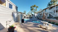 This outdoor communal space serves as a central gathering area within a residential complex, offering a well-maintained pool and an adjacent spa as its primary features. The pool is generously sized with clear, blue water and is accessible via built-in entry steps, while the circular spa sits nearby with its own tiled surround and jets. The surrounding deck provides ample room for lounge seating and relaxation under the open sky, benefiting from abundant natural sunlight throughout the day.

The complex is framed by two-story residential buildings with neutral-toned lap siding, balconies with metal railings, and exterior staircases. Mature eucalyptus and palm trees rise above the rooftops, adding a lush, California-casual aesthetic to the environment. Landscaped planting beds featuring ornamental grasses and seasonal blooms line the walkways, contributing to a tidy and welcoming atmosphere.

Additional amenities visible within the space include a utility/storage structure with French doors, decorative stone benches, potted plants, and a gated perimeter for privacy and security. The overall ambiance is relaxed and sun-drenched, characteristic of a well-kept Southern California residential community.