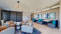 This contemporary single-floor amenity space makes a striking first impression with its polished concrete floors, high ceilings, and thoughtfully curated interiors. The main lobby entry features a dramatic textured wave wall, a suspended bubble chair, and a mounted display screen, establishing an immersive, design-forward atmosphere. Bold typographic wall art — including the words "IMMERSE" and "COMMUNICATE" — reinforces an intentional, community-focused identity throughout the space.

The expansive lounge area is anchored by a vibrant, multicolored area rug layered beneath mid-century modern armchairs and a low coffee table, illuminated by a sculptural multi-arm pendant light fixture. A slatted wood partition screen adds architectural depth while partially dividing zones. Adjacent to the lounge, teal and charcoal booth seating pairs with white-topped tables beneath large abstract artwork in aquatic tones, offering a social and collaborative gathering area.

The space also includes dedicated private meeting rooms, each with distinct color identities — one featuring vivid blue carpeting and teal accent walls with a round white table, another with deep purple walls and a bold floral canvas. Polished exposed ductwork and recessed lighting run throughout, blending an industrial edge with refined, gallery-quality finishes that elevate the overall ambiance.