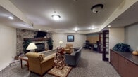 This single-floor community space is thoughtfully designed to provide comfort and functionality across several distinct areas. The centerpiece of the lounge is a striking floor-to-ceiling stone fireplace with a rustic wood mantel, wall-mounted television, and recessed lighting overhead. Comfortable seating arrangements, including upholstered armchairs and sofas set atop patterned area rugs, create an inviting atmosphere for relaxation and socializing. A dedicated computer workstation area is conveniently situated nearby, offering residents easy access to technology. Adjacent to the lounge, a bright dining area features multiple tables with seating for groups, set beneath decorative patterned valances framing large multi-pane windows and glass doors. These expansive windows and French doors flood the space with natural light and provide pleasant views of the surrounding landscaped grounds. The fully equipped kitchen includes dark cabinetry with brushed nickel hardware, granite-style countertops, a stainless steel refrigerator, microwave, and a double sink — all set on light tile flooring. Throughout the space, warm yellow accent walls, hardwood-style flooring in the dining area, and carefully chosen décor contribute to a cohesive, welcoming ambiance suited for communal living.