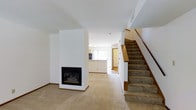 This two-story space is characterized by its clean, minimalist design and neutral color palette. The main level centers around an open living area finished with wall-to-wall carpet, creating a warm and comfortable atmosphere. A standout feature of the space is the floor-to-ceiling white fireplace surround, which serves as a striking architectural focal point and naturally divides the living area from the rest of the main floor. The fireplace itself is framed with a sleek black surround, adding a modern contrast to the bright white walls.

Beyond the fireplace, the space opens into an additional living or dining area, with a pass-through or open kitchen visible in the background, allowing natural light to flow through from a window. The layout provides a sense of connected, open-concept living on the main floor.

A carpeted staircase with a warm wood railing leads to the second floor, positioned along the right side of the space. The upper level overlooks the main floor, adding vertical dimension to the interior. Throughout, the white walls and light-toned carpet contribute to a bright, airy ambiance well-suited for comfortable everyday living.