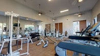 This single-floor space offers a thoughtfully designed environment that blends functional fitness amenities with refined interior living areas. The fitness center is generously sized, featuring high ceilings with elegant pendant lighting, large wall-to-wall mirrors that amplify the sense of space, and an abundance of natural light streaming through wood-slatted windows. The room is outfitted with a comprehensive selection of Precor and other branded equipment, including weight machines, a full dumbbell rack, benches, and cardio machines, all arranged on durable wood-patterned flooring. The interior living area presents a distinct artistic character, with crisp white walls serving as a gallery backdrop for large-scale framed artwork. A striking round green marble dining table with a pedestal base anchors the dining space, surrounded by woven rattan chairs and set atop a natural fiber rug. Concrete flooring adds an industrial edge throughout. A sliding barn door on a black rail system provides both a functional and decorative transition between spaces, while a vintage-style glass-paneled door connects to a hallway lined with additional framed artwork. The overall ambiance is sophisticated, blending contemporary design with curated artistic details.