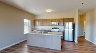 This single-floor residence is designed with a clean, contemporary aesthetic and a functional open-concept layout. The kitchen serves as a central feature of the living space, equipped with a full suite of stainless steel appliances, including a refrigerator, electric range, and built-in microwave. Warm-toned wood cabinetry provides generous storage, while granite-style countertops extend across both the perimeter workspace and a substantial peninsula island fitted with a double basin sink. The kitchen flows seamlessly into the main living area, which is finished with durable hardwood-style flooring that carries throughout the common spaces, enhancing the sense of continuity and openness. Natural light enters through multiple windows fitted with horizontal blinds. The living area features sliding glass doors that open to a private balcony with exterior views. The bedrooms are carpeted for comfort and include windows offering outdoor views. Bathrooms are finished with granite-topped vanities, wood-toned cabinetry, and tile-style flooring; one bathroom includes a walk-in shower, while another presents a half-bath configuration. Neutral wall tones throughout contribute to a cohesive, modern atmosphere.