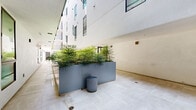 Located at 4971 S Centinela Ave in the Mar Vista neighborhood of Los Angeles, this ground-floor hallway and courtyard corridor offers a thoughtfully designed transitional space within a contemporary multi-story building. The space is defined by its clean, modern aesthetic, featuring light-toned stone tile flooring that runs the full length of the corridor, creating a sense of continuity and openness.

Large steel planter beds, finished in a muted slate blue, line the corridor and are filled with lush, tropical foliage that introduces a vibrant natural element against the crisp white stucco walls. Wooden benches are arranged along the planters, providing functional seating while complementing the organic tones of the greenery. The open-air courtyard design allows natural light to pour in from above, with upper-level balconies framed by blue railings visible overhead, adding vertical dimension and architectural interest.

Floor-to-ceiling glass sliding doors open into an interior common area, maintaining visual connectivity between the indoor and outdoor spaces. The multi-story building facade, with its grid of windows, rises prominently on one side, contributing to a sheltered yet airy atmosphere. The overall ambiance is calm, clean, and welcoming — a well-considered passageway that balances modern design with natural elements.