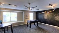 This single-floor space is designed as a dedicated recreation and activity room, combining functional design with a fun, engaging atmosphere. The room features neutral gray walls complemented by a full chalkboard accent wall, which serves as both a creative and decorative element, adorned with colorful bunting and handwritten notes. The chalkboard wall adds a dynamic, interactive quality to the space.

The room is carpeted with a multi-toned gray carpet tile, providing a durable and comfortable surface underfoot. Natural light enters through multiple windows fitted with blinds, keeping the space bright while allowing for light control. A large ceiling fan with an integrated light kit and accompanying track lighting ensure the area remains well-lit at all times.

A full-size foosball table serves as the centerpiece of the room, making it an ideal setting for recreational activities and social gatherings. Small wall-mounted book ledges add a practical touch, offering storage for books or small items. The overall layout provides ample open floor space around the foosball table, allowing for easy movement and additional activities. The space conveys a lively, welcoming ambiance suited for both children and adults.
