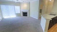 This single-floor residence offers a well-appointed living area defined by classic architectural details and a functional open layout. The main living space is finished with plush wall-to-wall carpeting in a neutral beige tone, creating a warm and comfortable foundation throughout the room. Crown molding lines the upper perimeter, adding a refined architectural touch to the otherwise clean, softly painted walls.

A white-mantled fireplace serves as the room's focal point, positioned along the central wall and flanked by generous open floor space on either side. A ceiling fan with a light kit is centrally mounted, providing both comfort and ambient illumination, while a track lighting fixture adds additional directed light. Vertical blinds on a large window allow for adjustable natural light, brightening the interior throughout the day.

To one side, a partial wall with a light stone countertop hints at an adjacent kitchen area, suggesting an open or semi-open floor plan that promotes easy flow between spaces. A hallway visible in the background indicates further rooms beyond the main living area. Overall, the space presents a clean, move-in-ready environment with classic detailing and a practical, connected layout.