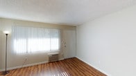 This single-floor space offers a clean, straightforward layout designed for comfortable everyday living. The main living area is finished with warm, wood-toned laminate flooring that adds a sense of warmth and continuity throughout the space. A large window fitted with vertical blinds allows an abundance of natural light to flow in, creating a bright and welcoming atmosphere. The textured popcorn ceiling adds a classic, familiar character to the room. A wall-mounted air conditioning unit ensures year-round climate comfort, positioned conveniently beneath the window. The neutral, light-toned walls provide a versatile backdrop that easily accommodates a variety of interior styles and décor preferences. A solid entry door with a standard lock offers straightforward access and a sense of security. White baseboards frame the perimeter of the room, providing a crisp finishing detail that ties the space together. Overall, the space presents a functional and practical environment with essential amenities, making it well-suited for those seeking a simple, low-maintenance living arrangement.
