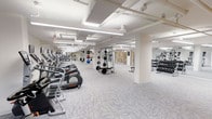 This fitness center offers a comprehensive workout environment designed to accommodate a variety of exercise needs. The space is generously proportioned, with a clean, open layout that allows for easy navigation between different workout zones. Bright, recessed fluorescent lighting illuminates the entire floor, complemented by a crisp white color palette that keeps the atmosphere feeling fresh and energizing.

Along one side of the facility, a long row of cardio equipment — including elliptical trainers and treadmills — lines the wall, providing ample options for aerobic exercise. Toward the center and rear of the space, a dedicated strength training area features a multi-function cable and weight machine, along with a rack of medicine balls for functional fitness. A separate free weights section houses an extensive dumbbell rack, offering a full range of weight options for resistance training.

The flooring throughout consists of durable, patterned fitness carpet tiles that provide both comfort and practicality. Structural columns are integrated seamlessly into the layout without disrupting the flow of the space. Mirrored walls in key sections enhance the sense of openness while allowing users to monitor their form. The overall ambiance is clean, professional, and purpose-built for an effective fitness experience.
